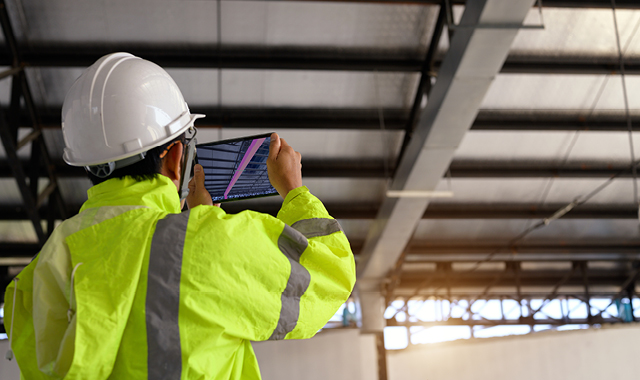 A man in white hard hat uses a tablet to take infrared imaging of ceiling ducts inside a warehouse.